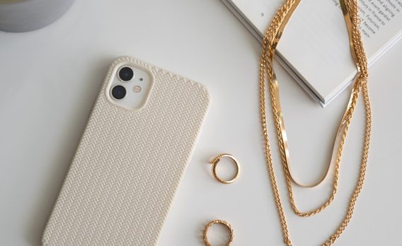 A cream phone case, two gold rings and layered gold chains on a white surface